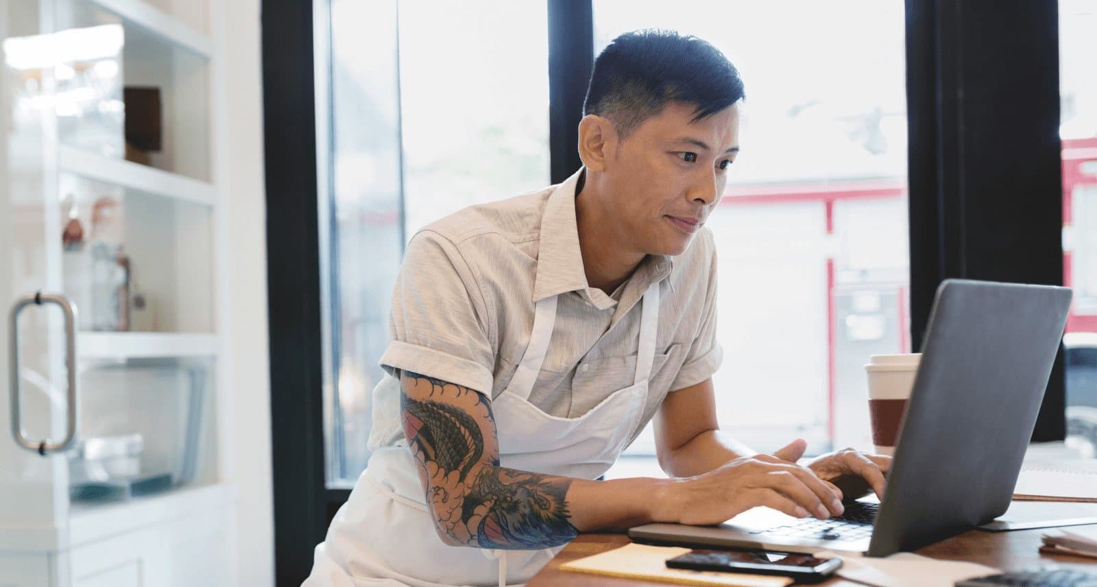 Young worker wearing an apron with tattoo sleeve using a laptop